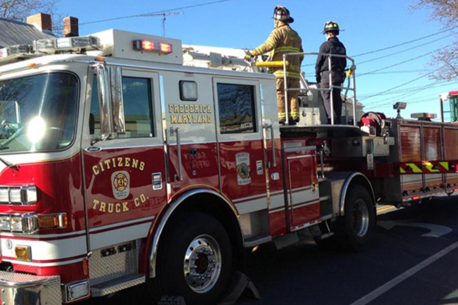 Two firefighters stand on top of a red and white Citizens Truck Co. ladder truck from Frederick, Maryland, parked on a sunlit street.