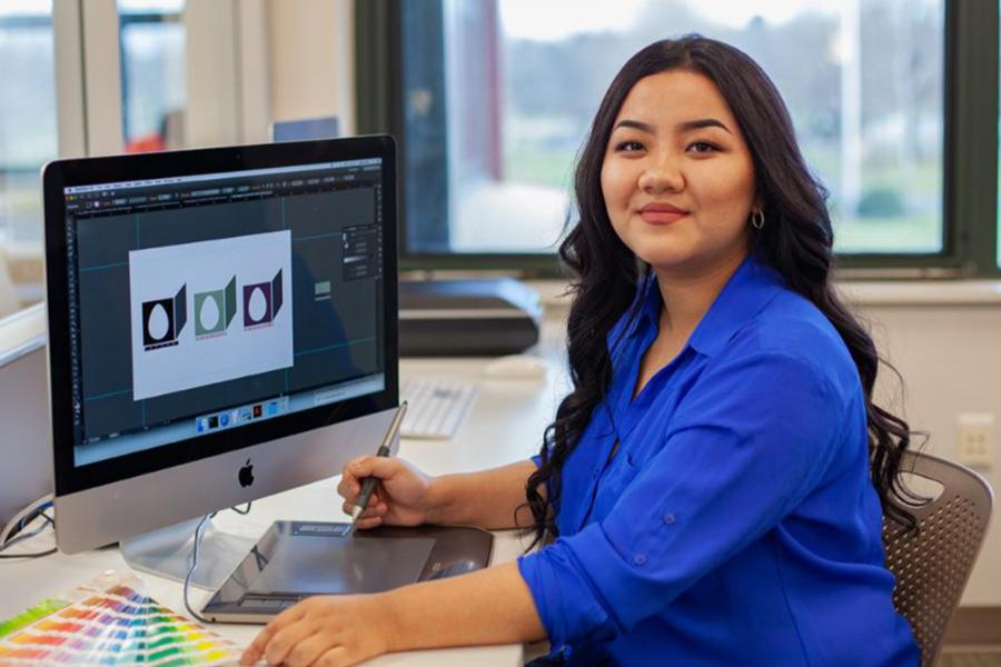 A young woman in a blue shirt smiling at her desk while using a stylus and graphics tablet to design a logo on an iMac.