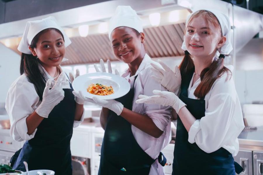 Three smiling young female culinary students in chef hats, black aprons, and white gloves proudly presenting a finished pasta dish in a commercial kitchen.