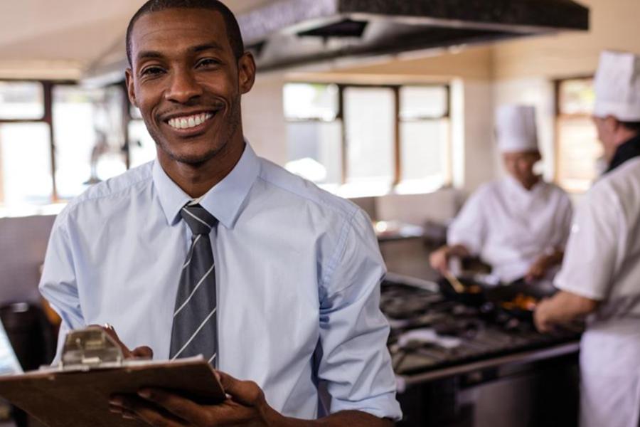 A smiling man in a light blue button-down shirt and grey tie holding a clipboard in a professional kitchen, with chefs working in the background.