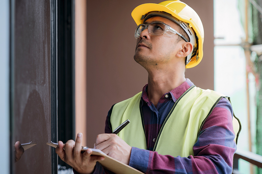 A home inspector wearing a yellow hard hat, safety glasses, and a high-visibility vest takes notes on a clipboard while examining the exterior of a building.