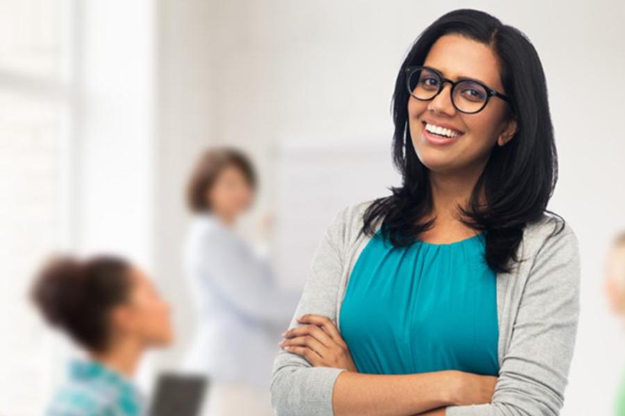 A professional portrait of a woman in a teal top and grey cardigan, smiling with arms crossed in a bright, modern office or campus setting.