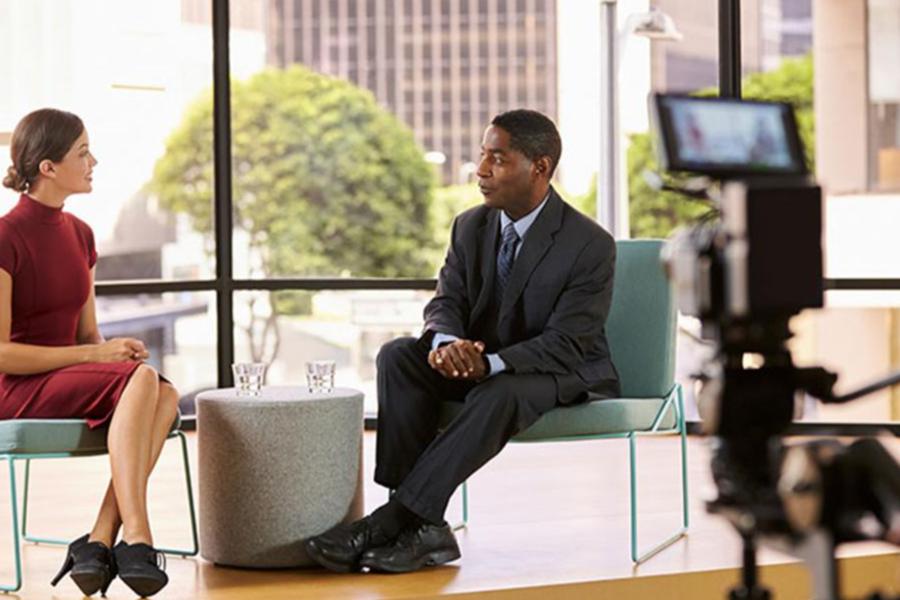 A male and female professional participating in a televised interview or talk show in a modern studio with a city view.