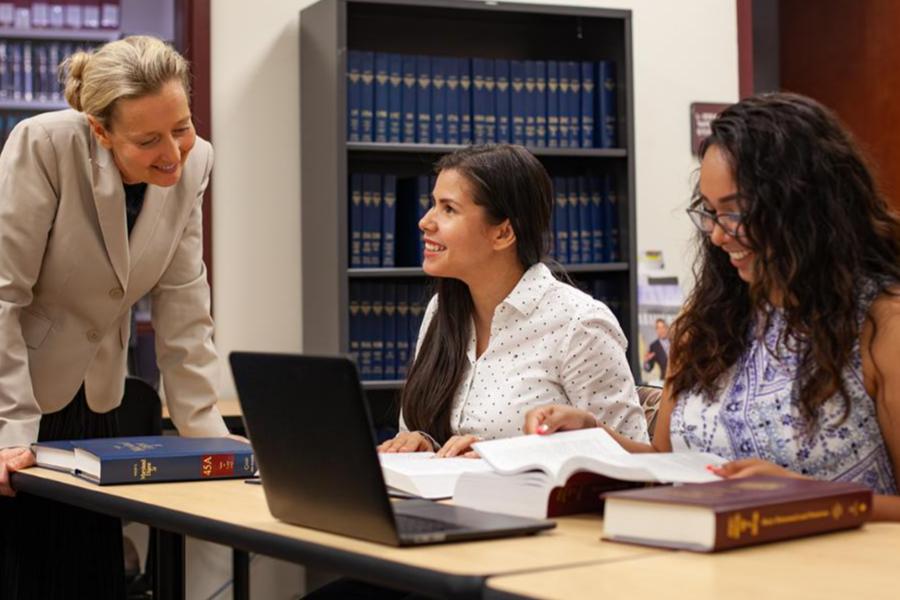 A librarian or instructor in a beige blazer leans over a table to assist two students who are researching with open law books and a laptop in a library with tall bookshelves.