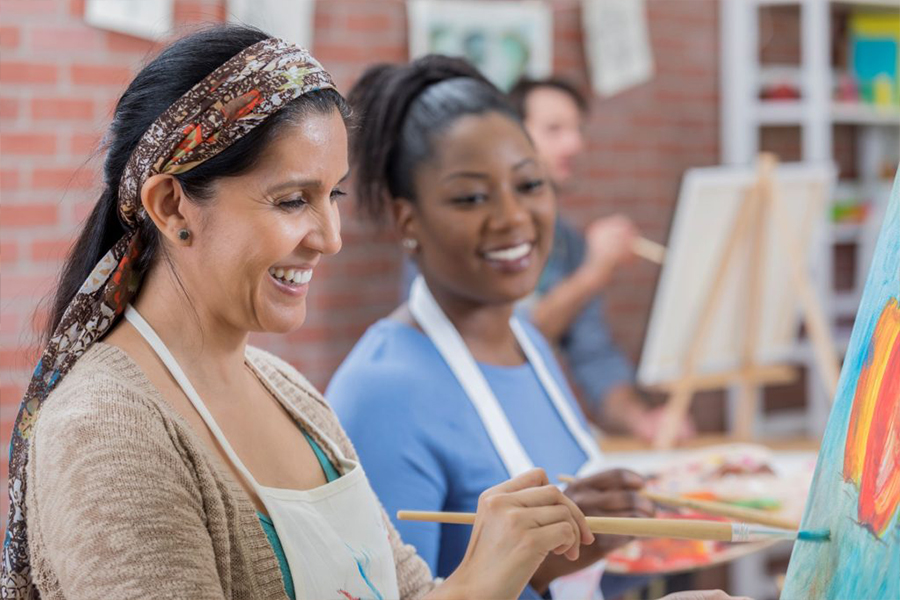 Two women smile while painting on canvases at easels in an art class, with other students working in the background.