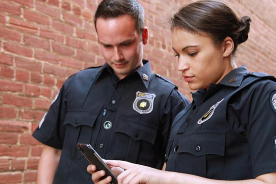 Two police officers in dark uniforms stand together in front of a brick wall, both looking down at a smartphone. They appear focused on the screen, representing teamwork, digital communication, and professional development in the field of public safety.