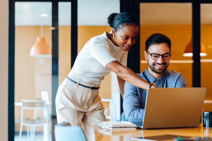 Two smiling colleagues, a woman and a man, collaborating on a project over a laptop in a modern office.