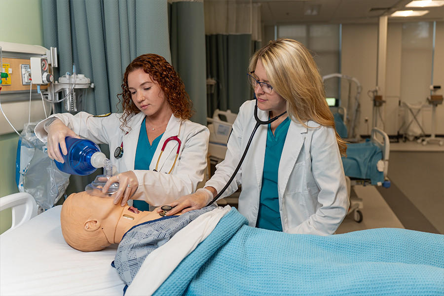 Two Respiratory Care students in white lab coats and teal scrubs work together on a medical simulation mannequin in a clinical training lab. One student administers manual ventilation using a bag-valve mask while the other uses a stethoscope to assess breath sounds.