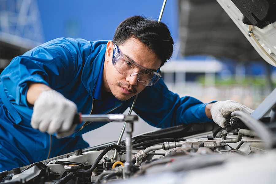 An automotive technician in a blue uniform and safety glasses uses a wrench to work on a car engine under an open hood.