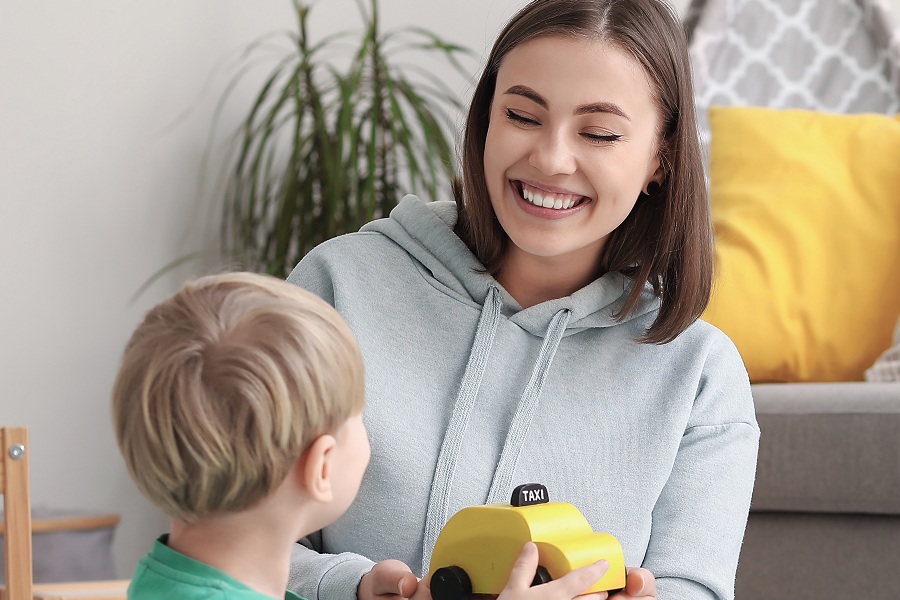 A smiling young woman plays with a toddler on the floor of a living room, both holding a yellow wooden toy taxi cab. A couch with a yellow pillow and a houseplant are visible in the background.