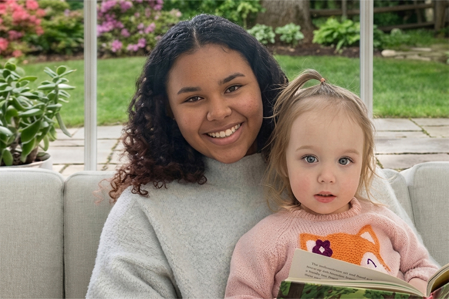 A smiling teenage girl sits on a couch with a toddler in a pink fox sweater, who holds an open book. A garden with flowering plants is visible through the window behind them.