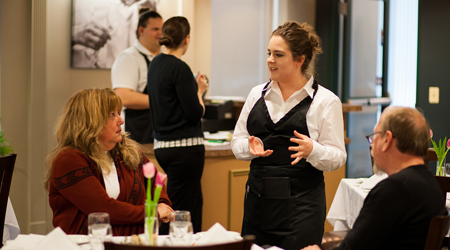 A server in a white shirt and black apron stands at a table with two customers, explaining a menu in a fine-dining setting.