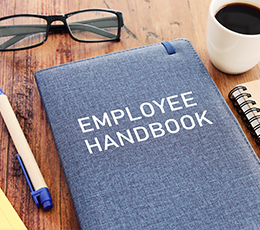 An Employee Handbook on a wooden desk with coffee and office supplies.