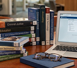 A stack of college textbooks beside an open laptop and glasses on a desk.