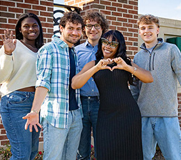 Five smiling college students pose together outdoors in front of a brick building, with one making a heart shape with her hands.