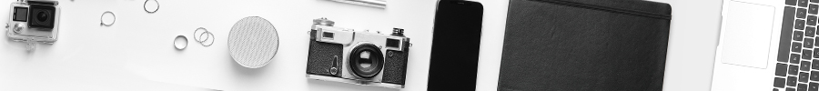 Photography and technology equipment laid out on a white surface, including a vintage film camera, GoPro action camera, smartphone, portable speaker, leather notebook, and laptop.