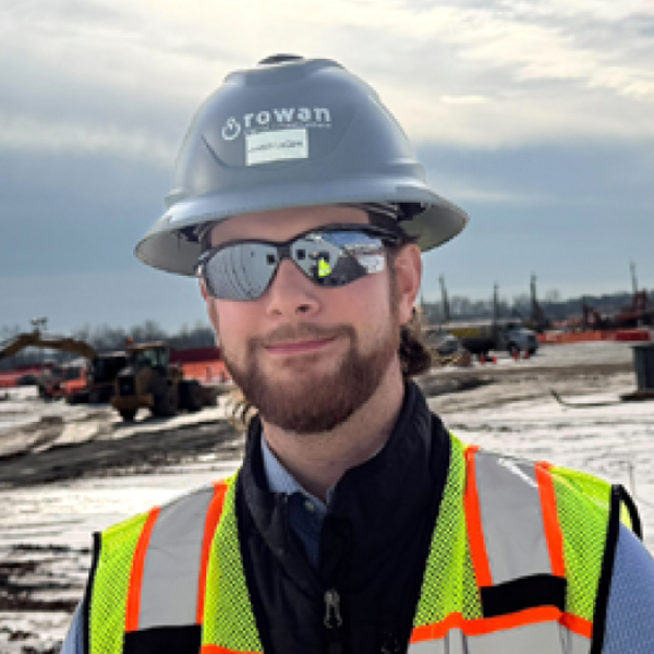A man wearing a hard hat, safety vest, and sunglasses stands at a construction site with machinery and piles of dirt in the background.