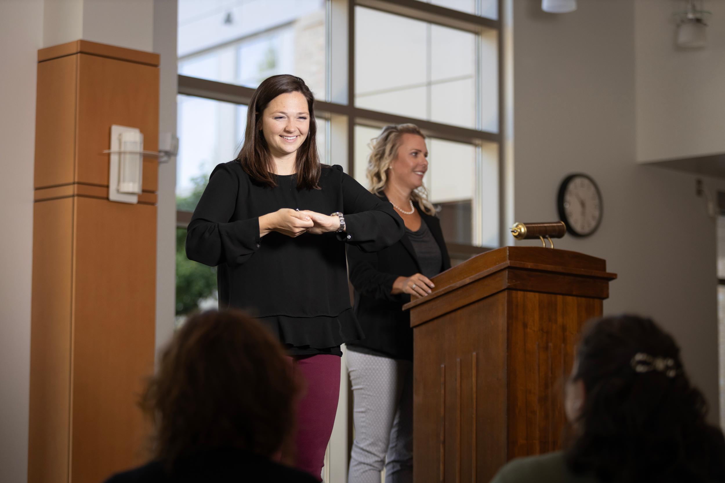Woman wearing black top signing to 2 people. Another woman is speaking at a podium.
