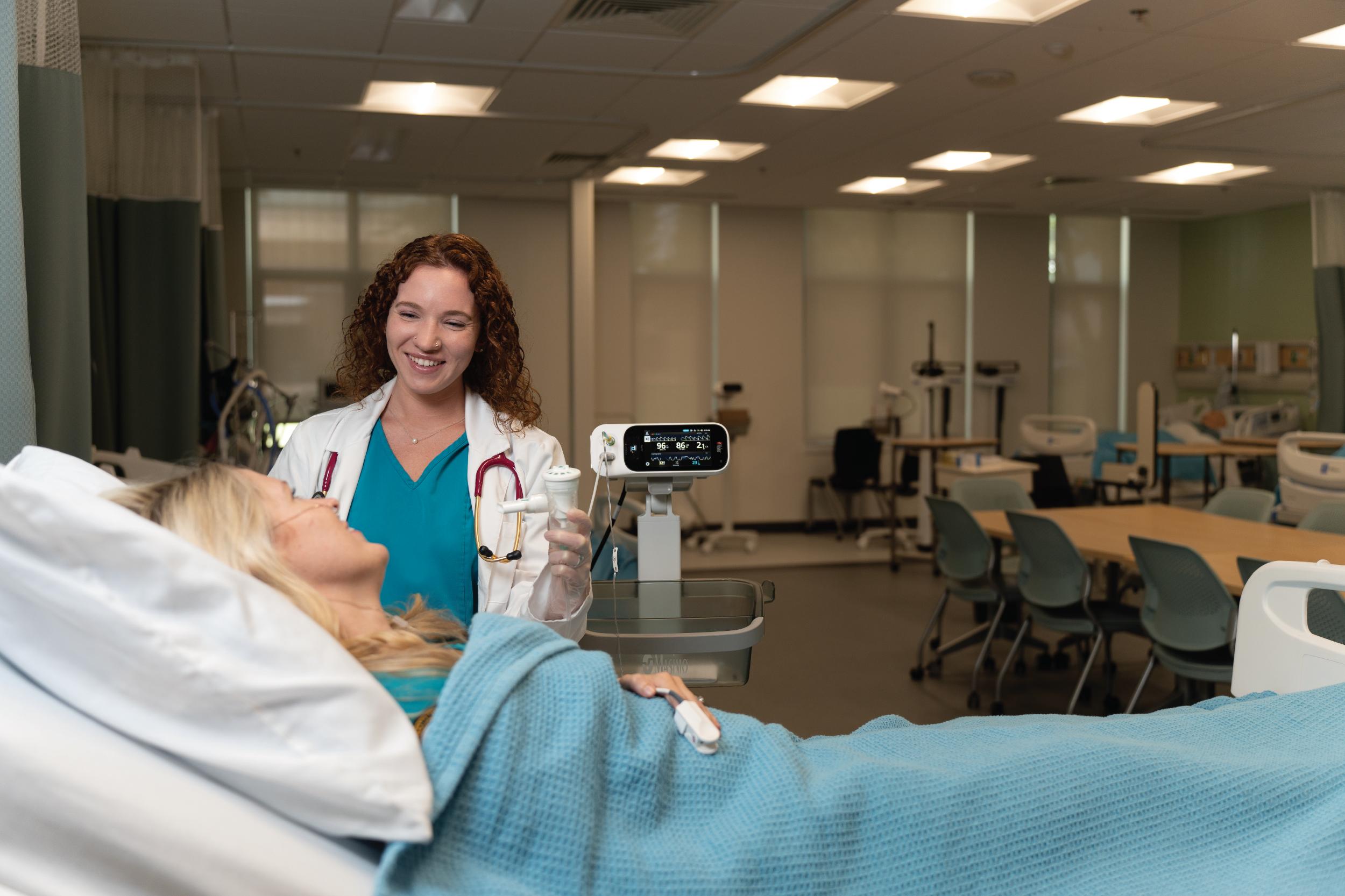 A woman in turquoise scrubs and white lab coat talking with a female patient in a hospital bed.