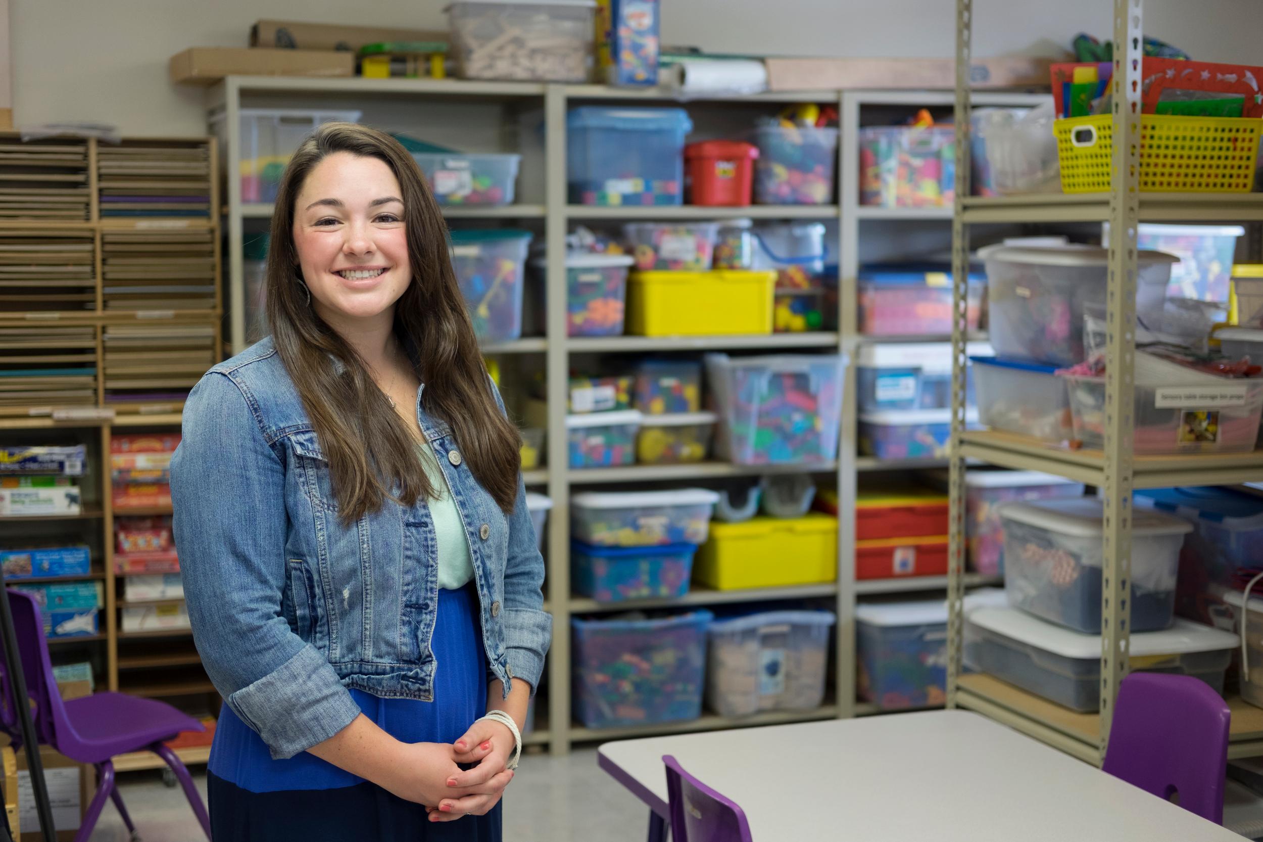 woman in denim jacket stands in front of shelves with classroom supplies
