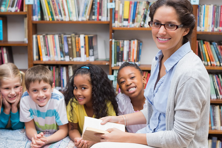 A smiling teacher holds an open book while seated with four young children in a colorful school library.