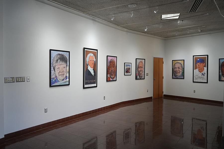 An art gallery features white walls lined with several framed portrait paintings under professional track lighting. The polished floor reflects the artwork, and a wooden door is visible at the end of the long wall.