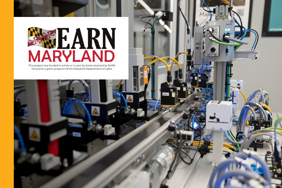 Complex industrial machinery with robotic components and yellow wiring is shown in a laboratory or factory setting. A white overlay in the corner features the EARN MARYLAND logo and a statement noting the project was funded by the Maryland Department of Labor.