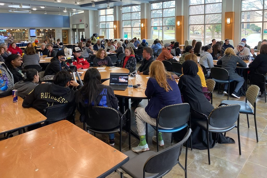 A large group of people are seated at round tables in a bright, spacious cafeteria or community room with large windows. They are engaged in discussions, with some tables featuring laptops and cameras