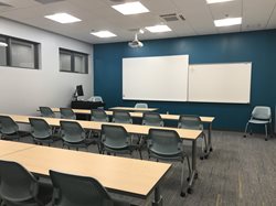 A modern, empty classroom with several rows of long wooden tables and grey chairs. A projector is mounted to the ceiling facing two large whiteboards on a dark teal accent wall.